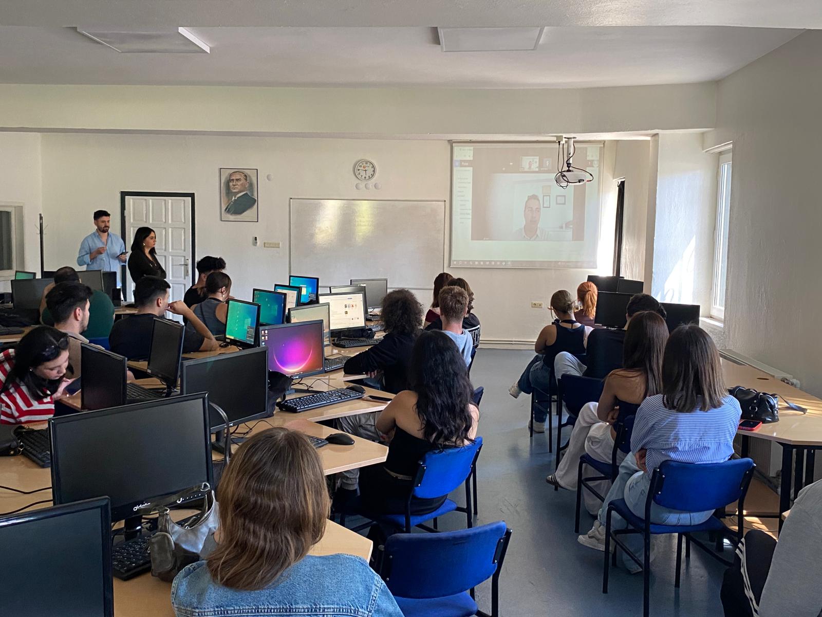 In a computer lab, approximately 20–25 university students are watching the image projected on the screen. The lab is simple, featuring white walls, light-colored desks, and rectangular LED lights on the ceiling. The students are mostly young adults, and most appear to be seated at the desks, focused on the lesson.